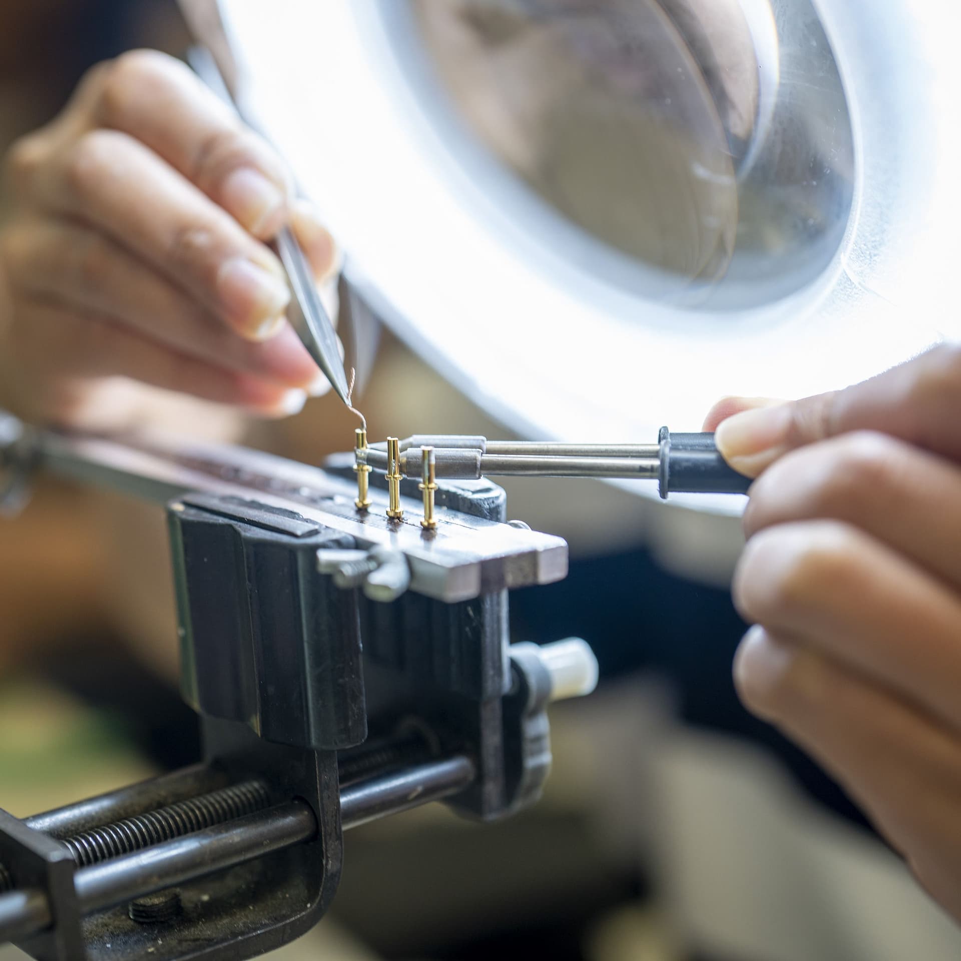 Hands holding a wire with tweezers over terminal posts clamped in a bench vise, under a magnifier lamp.