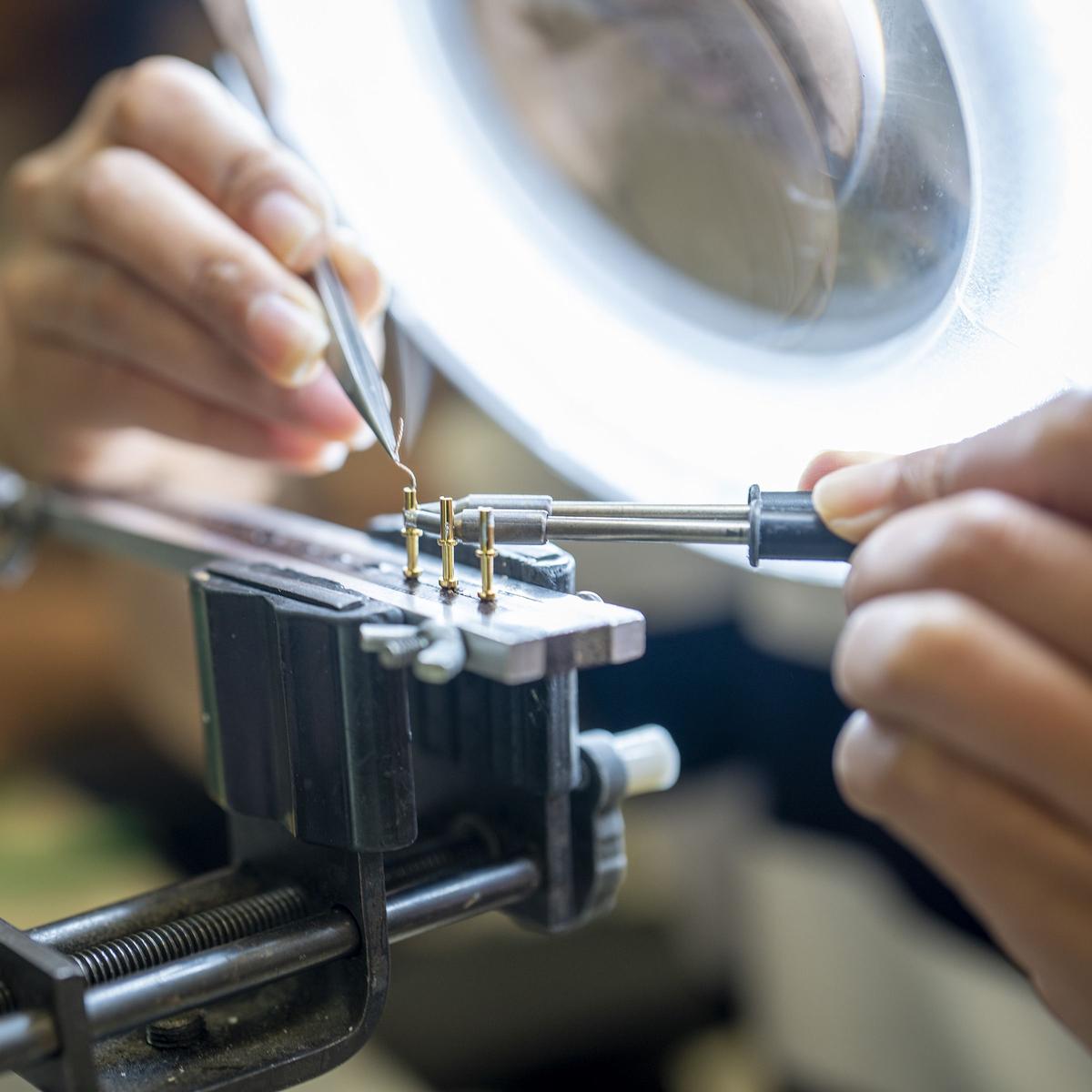 Hands holding a wire with tweezers over terminal posts clamped in a bench vise, under a magnifier lamp.