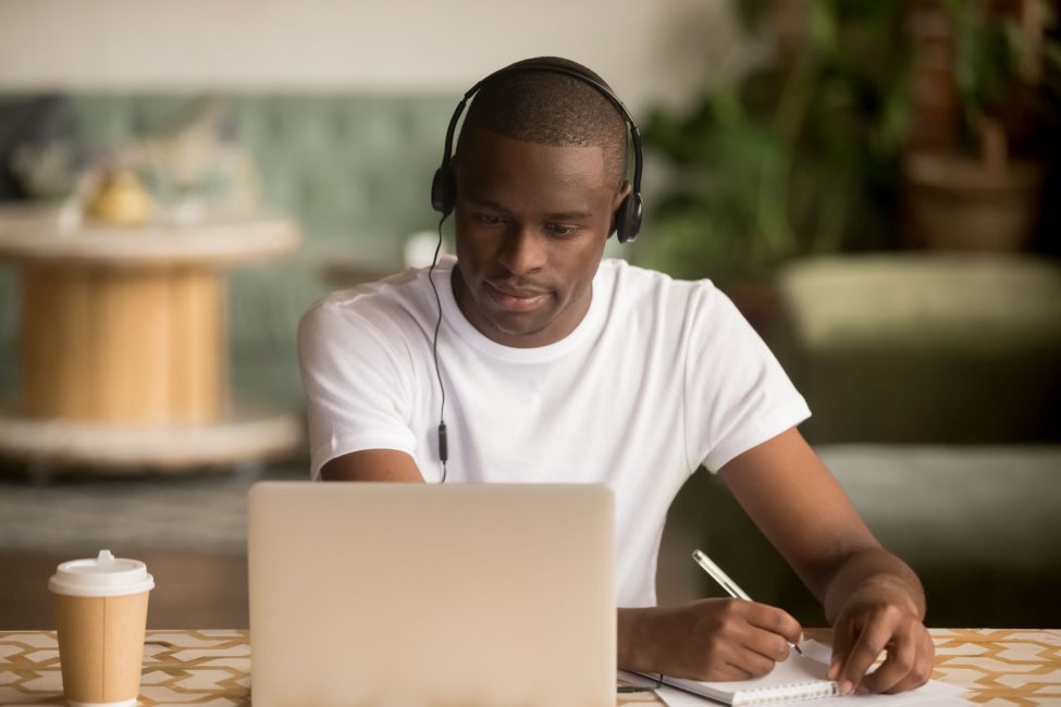 Focused african man wearing headphones watching webinar training making notes study online learning language on computer, black male student looking at laptop elearning in internet write information